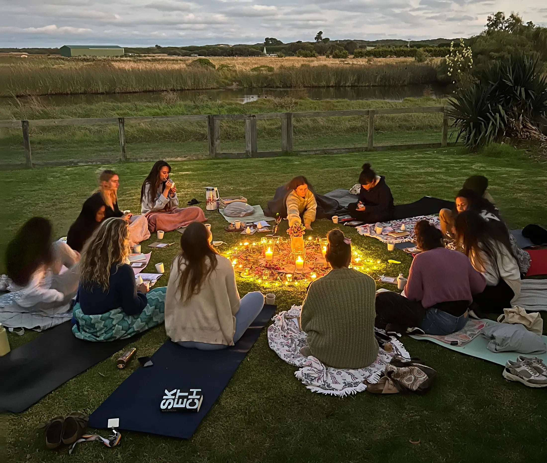Un círculo de mujeres sentadas en el pasto al atardecer, alrededor de un altar de velas encendidas.