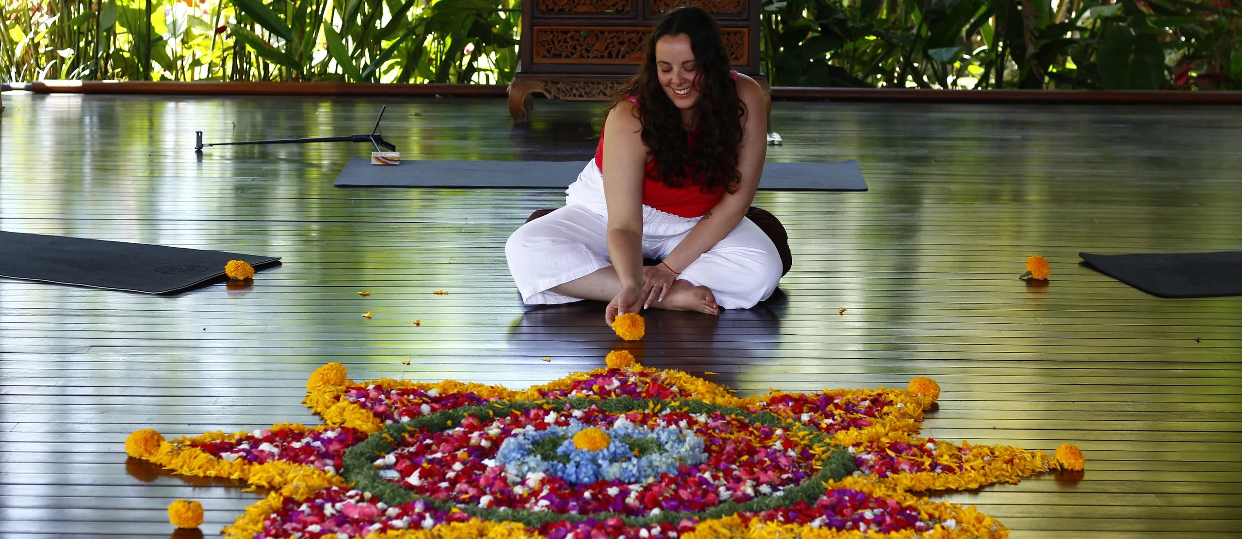 Marisa sentada en una terraza de madera, colocando flores en un mandala grande, rodeada de vegetación.